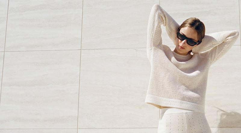 A model wearing a cream knit outfit against a pale stone wall
